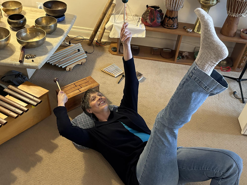 Person lying on a cushion, smiling, raising mallets with instruments around. Cozy room, beige carpet, and colorful percussion instruments.
