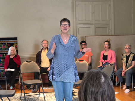 A person speaks to a small seated group in a room with a patterned tapestry and neutral walls. The mood is engaging and attentive.