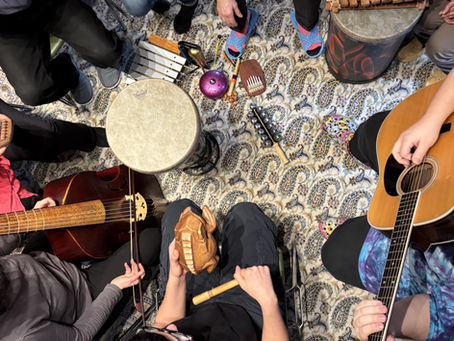 A group of people play various instruments, like drums and guitar, in a circle on a patterned rug. Some wear colorful socks, creating a lively vibe.