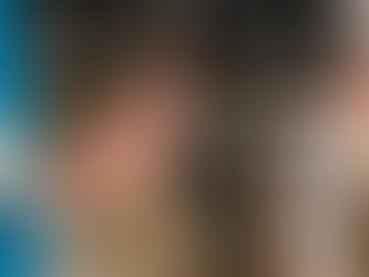 Two people seated indoors, one holding a drum and drumsticks, the other an instrument. Wooden floor, amplifier in background.