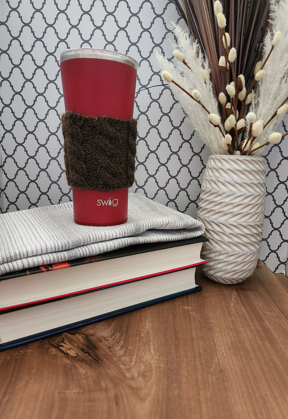 Thumbnail: Brown cabled knit coffee cozy on a red coffee tumbler travel mug posed on grey/white kitchen towel