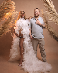 Pregnant couple posing for a maternity portrait in Reading, PA. The wife is wearing a white tulle maternity gown with her belly exposed the husband is in a white button up with beige pants. They are surrounded by pampas grass
