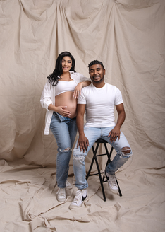 husband and wife posing for portraits, they are both wearing jeans and a white tee shirt, he sits on a stool and she is standing beside him with on hand resting on the top of her belly