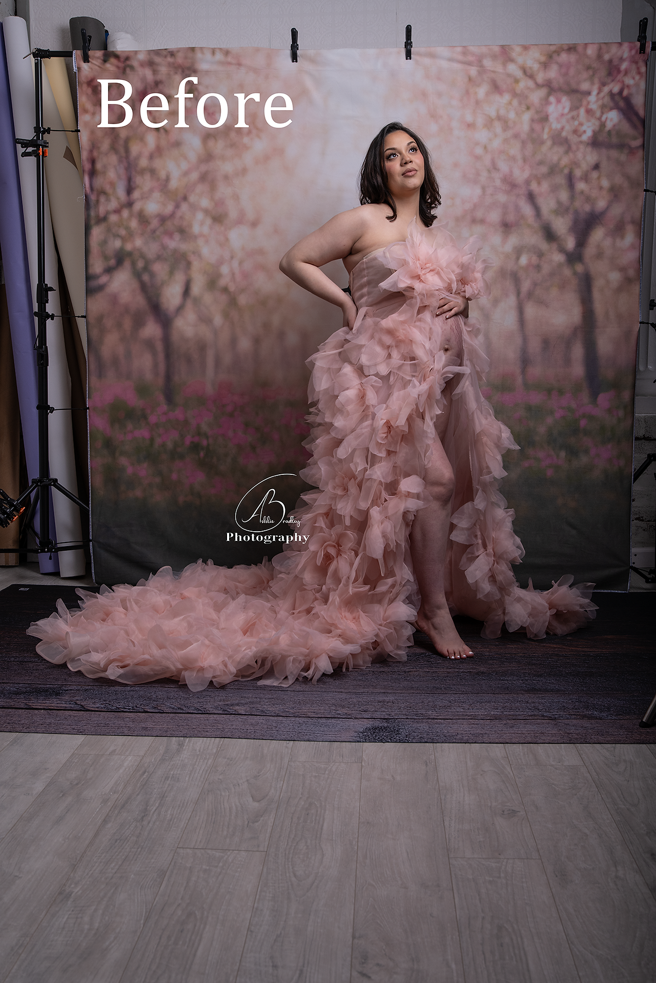 a picture of a maternity portrait before it is edited, the woman is wearing a light pink fluffy tulle down with her stomach exposed, she has one hand resting on her stomach and the other hand on the small of her back, she appears to be looking off into the corner. The background is a fabric backdrop of a cherry blossom orchard but isn't fully extended to the corner of the frame