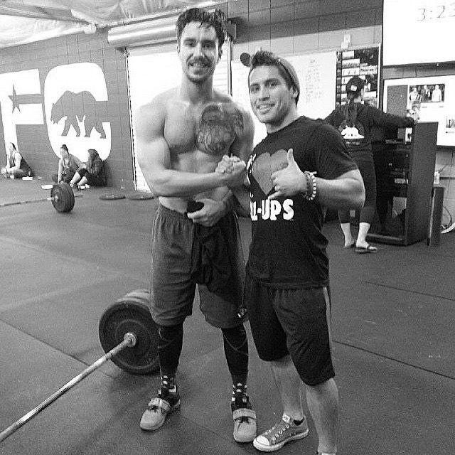 Two well muscled men shaking hands in the gym, one without his shirt on, the other with his shirt on while a several barbells are on the floor in the background