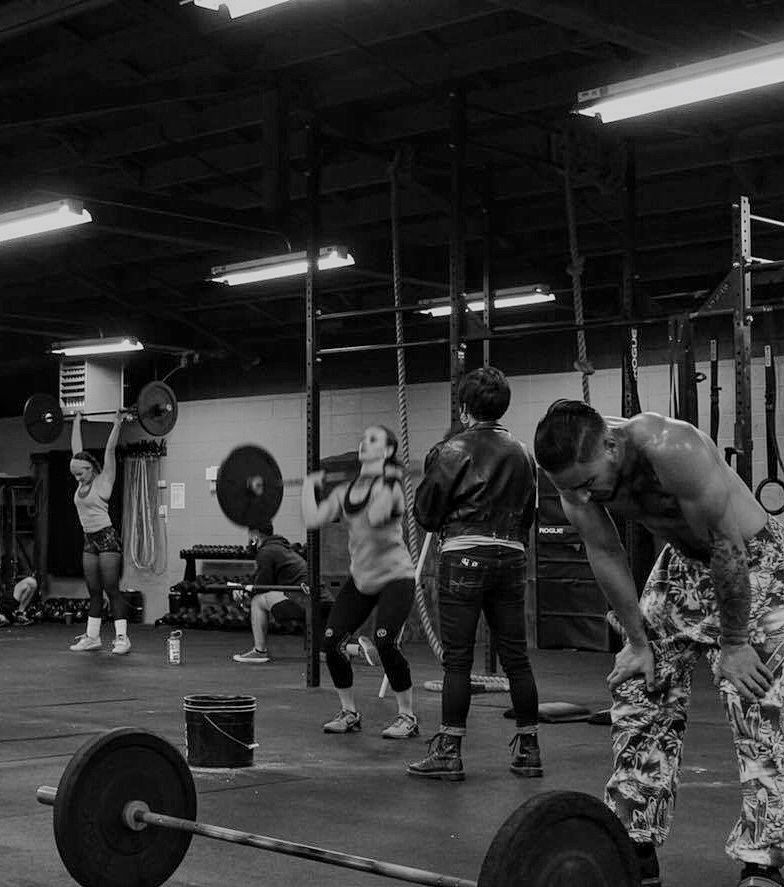 Well muscled man rests during a workout in the gym with his hands on his knees. There are other athletes in the back lifting barbells as he stands in front of his barbell on the ground