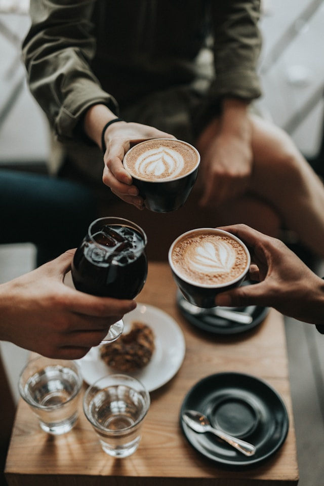 Three people toast with cappuccinos and iced coffee over a wooden table. Two saucers, glasses, and a cookie are visible. Cozy atmosphere.
Photo by Nathan Dumlao on Unsplash.