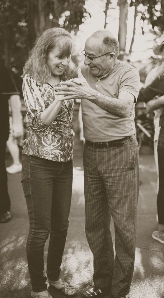An older couple dances happily outdoors, smiling warmly. The man wears a polo and stripes; the woman, a patterned blouse. Sepia-toned image. Via unsplash.com