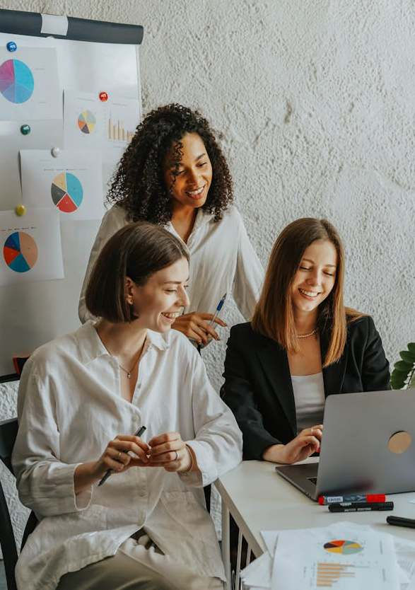 Three women in an office setting, smiling while discussing a laptop screen. Charts on the wall. Bright room, collaborative mood.