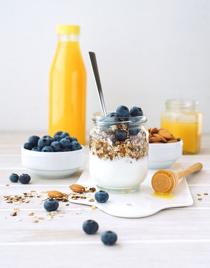 Yogurt jar with granola and blueberries, spoon inside. Surrounding white bowls with blueberries, almonds, orange juice bottle, honey dipper.