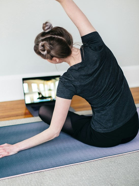 Woman doing yoga on a mat, stretching arm, hair in buns. Laptop nearby shows yoga video. Calm home setting, wood floor, gray wall.