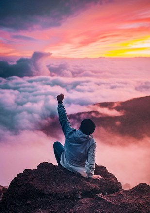 Person in a gray hoodie sits on a rock, raising a fist triumphantly. Sunset sky and clouds create a vibrant backdrop. Mood: victorious.