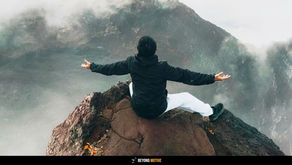 Person in black jacket sits on a rocky cliff with arms outstretched, surrounded by misty mountains. Text reads "Beyond Motive." Serene mood. How Living in the Present Moment Can Transform Your Life