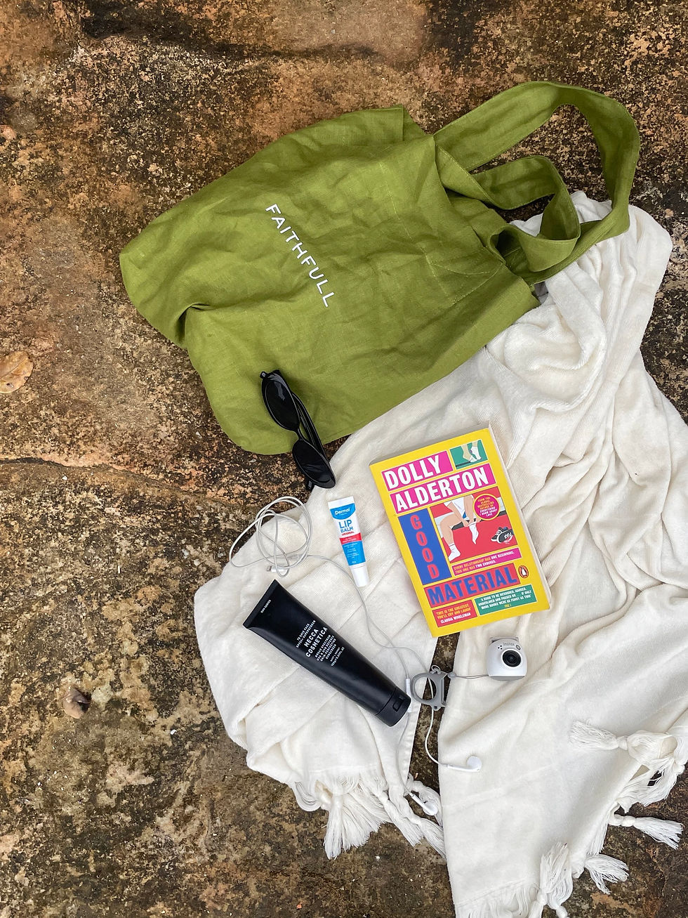 Green tote bag, cream towel, black sunnies, black packaged sunglasses, white wire headphones and the book Good Material by Dolly Alderton on a rock.