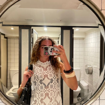 Woman taking a selfie in a bathroom mirror, wearing a patterned white top. The setting has tiled walls with a visible toilet.