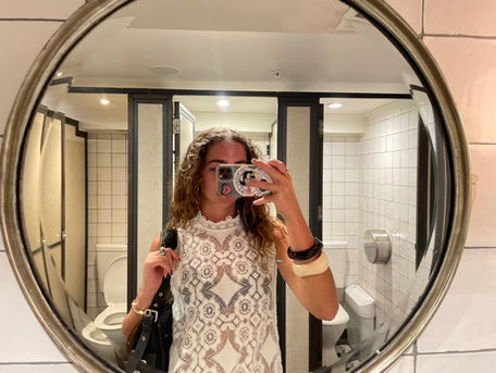 Woman taking a selfie in a bathroom mirror, wearing a patterned white top. The setting has tiled walls with a visible toilet.