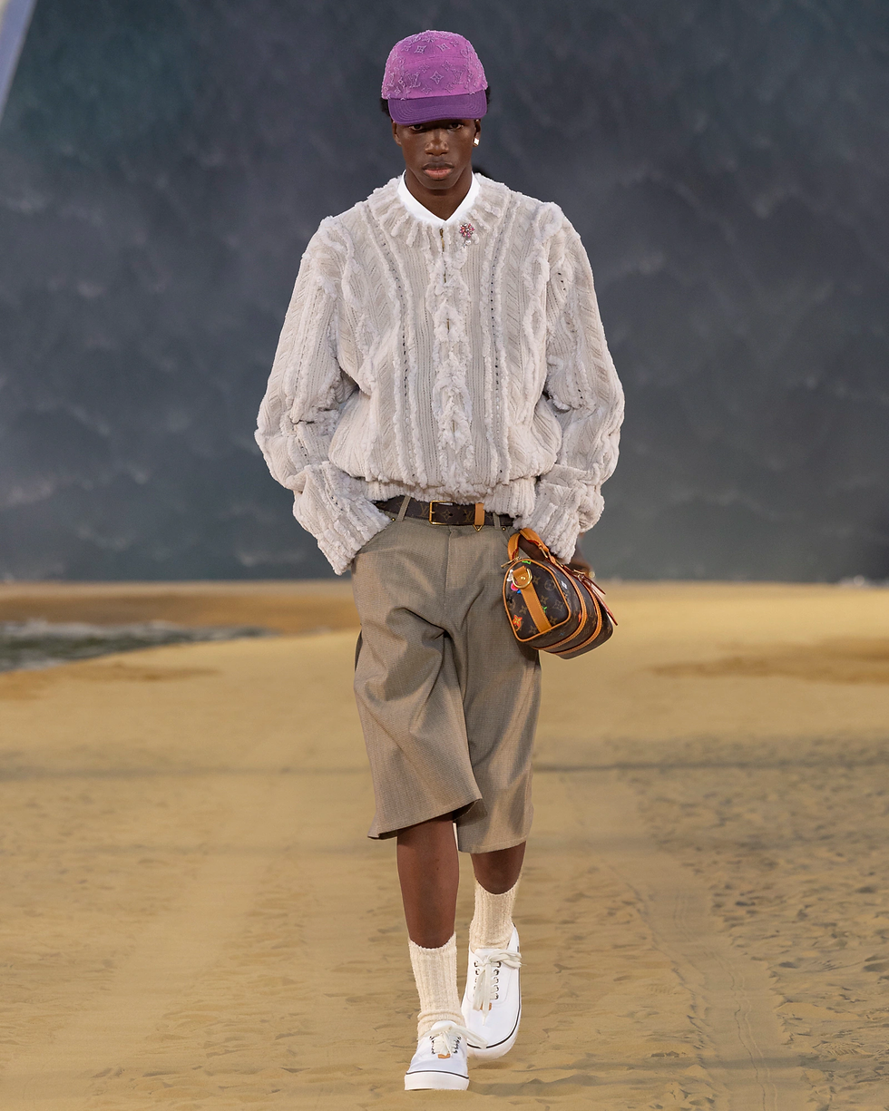 Runway model in purple cap and oversized cream knit, beige shorts, carrying a monogram bag, walking on a sandy stage.