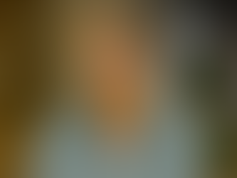 Smiling woman with short blonde hair wearing a light blue blouse, seated indoors with soft natural lighting and blurred bookshelves and plants in the background