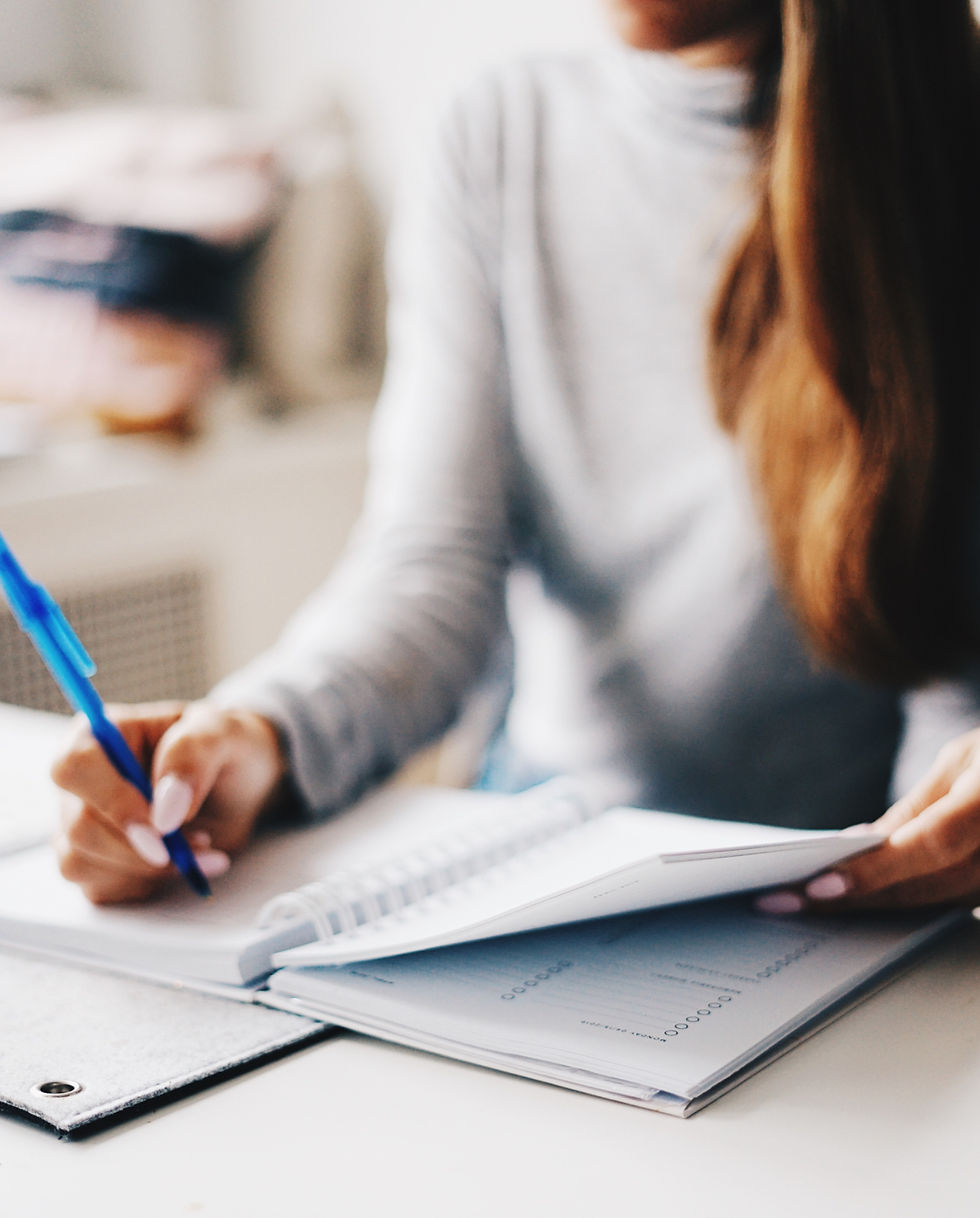Person writing with a blue pen in an open notebook beside a planner on a desk, in a softly blurred indoor setting.