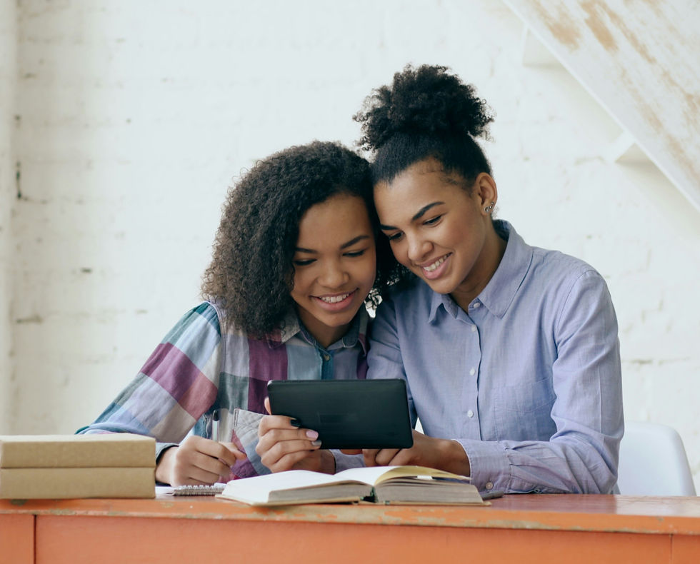 Two young women smile together over a tablet at a desk with books and boxes, in a bright white room, looking focused and happy
