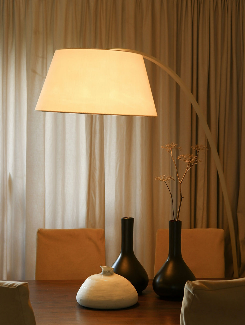 Warm dining room with a lit arched floor lamp, wooden table, black and white vases, beige chairs, and curtains