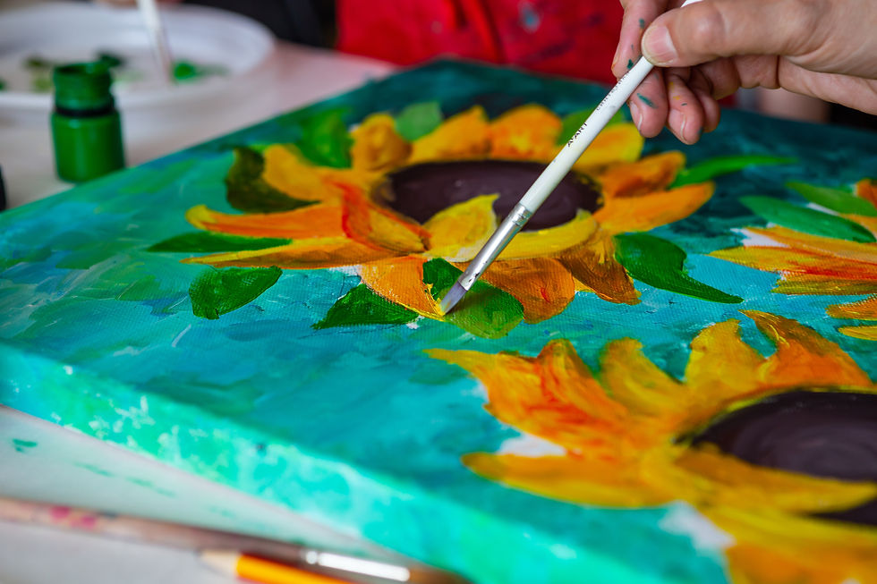 Hand paints a bright sunflower on a teal canvas with a brush, beside green paint jars.