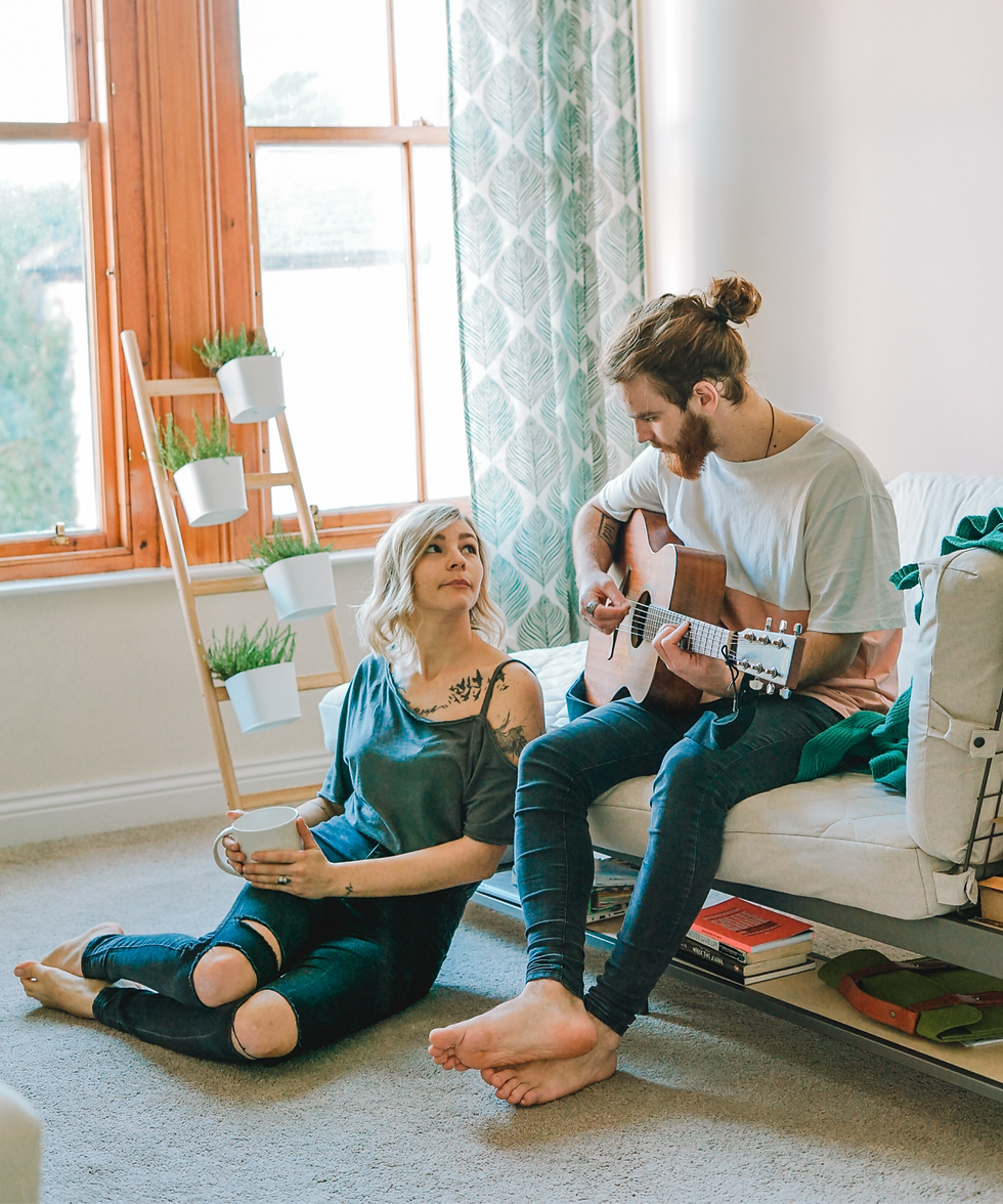 Woman with mug watches man play guitar in a bright living room with plants, window, and patterned curtain.