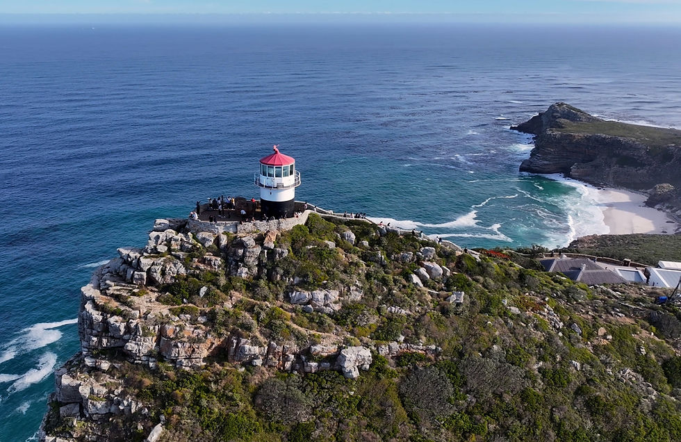 Cape Point Lighthouse. Red-and-white lighthouse on a rocky cliff above blue ocean, with visitors on the lookout and a sandy cove nearby.