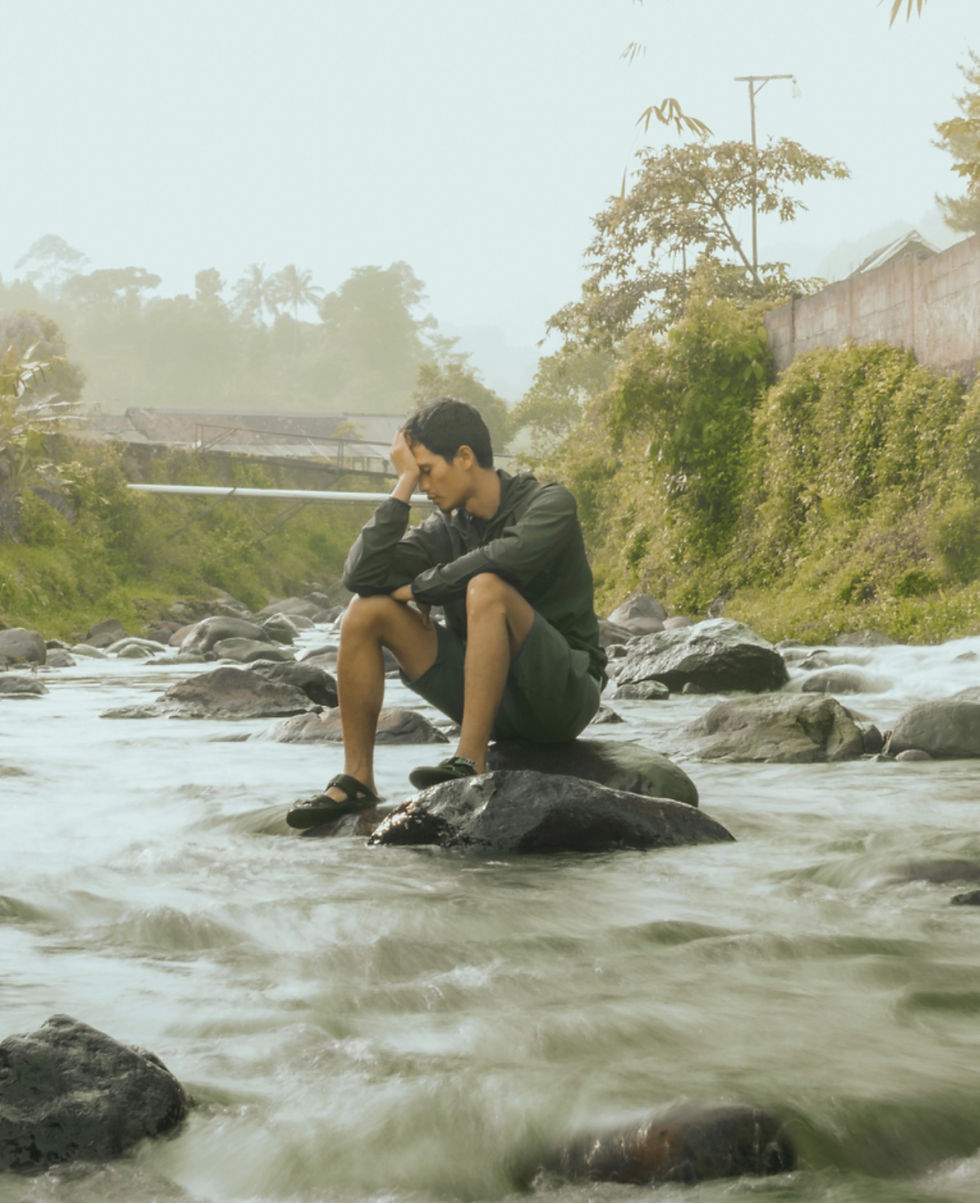 Man in dark clothes sits on a rock in a rushing river, head in hand, looking pensive amid misty green trees.