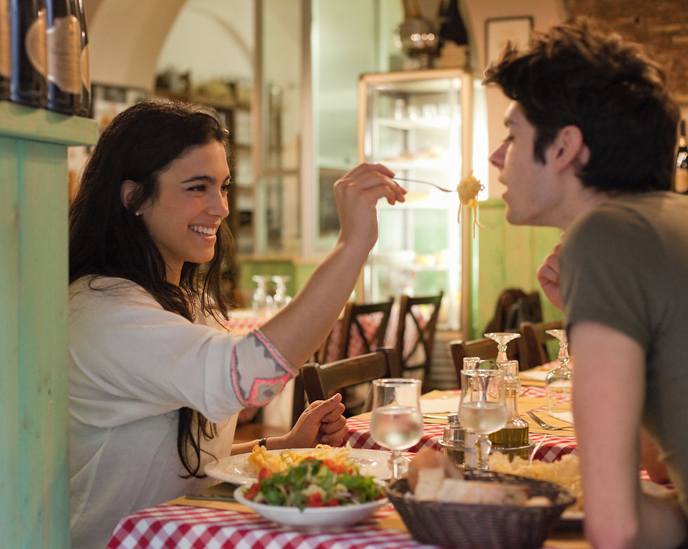 Smiling woman feeds pasta to a young man at a cozy restaurant table with red-checkered cloth, wine glasses, and salad.