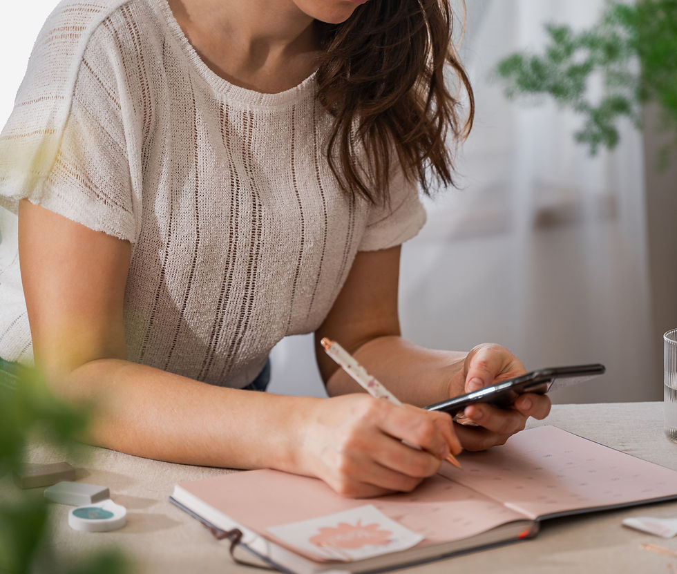 Woman in a cream sweater writes in a pink notebook while checking her phone at a bright desk with plants.