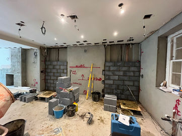 Interior construction site with unfinished walls and ceiling. The ceiling has multiple cutouts with exposed wiring and hanging light fixtures. Two partial block walls are being built against the back wall, with gray concrete blocks stacked nearby. Red markings and notes are written on the walls, indicating measurements and instructions. The floor is covered with construction materials, including buckets, tools, a level, and a blue equipment case. A window on the right side provides natural light. The overall space appears to be in the middle of a renovation or structural modification.