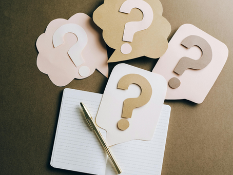 Open notebook with pen and four pastel speech bubbles with question marks on a brown background, suggesting a brainstorming mood