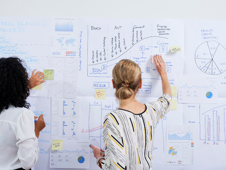 Two people standing in front of a wall covered with charts, graphs, sticky notes, and planning documents while reviewing and discussing the information.