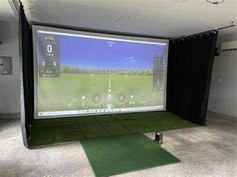 Golf simulator setup indoors with a large screen showing a virtual golf course. Green mat and projector are visible, creating a realistic scene.