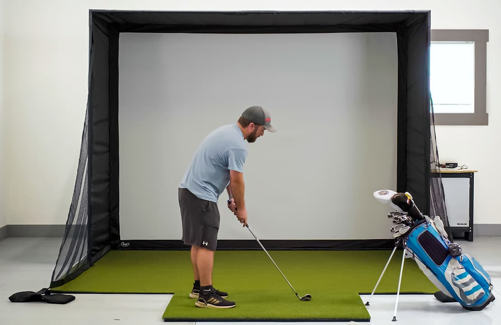 Man in a blue shirt practicing golf indoors on green turf, swinging a club, with a golf bag nearby and a large golf simulator screen behind.