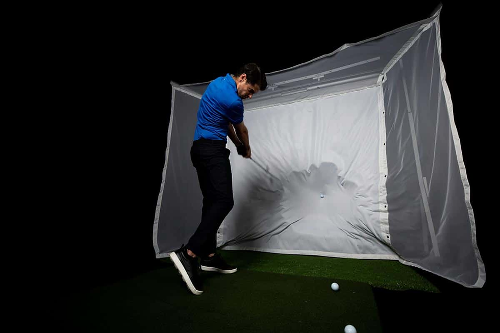 Man in a blue shirt swings a golf club, hitting a ball into a white net enclosure. Dark background, focused action, and concentration.