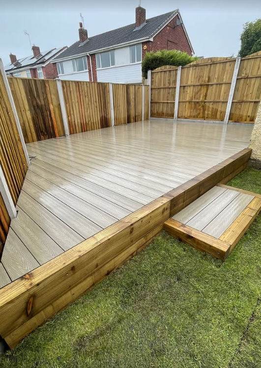 Light brown composite decking installation with raised platform and wooden step in a fenced garden.