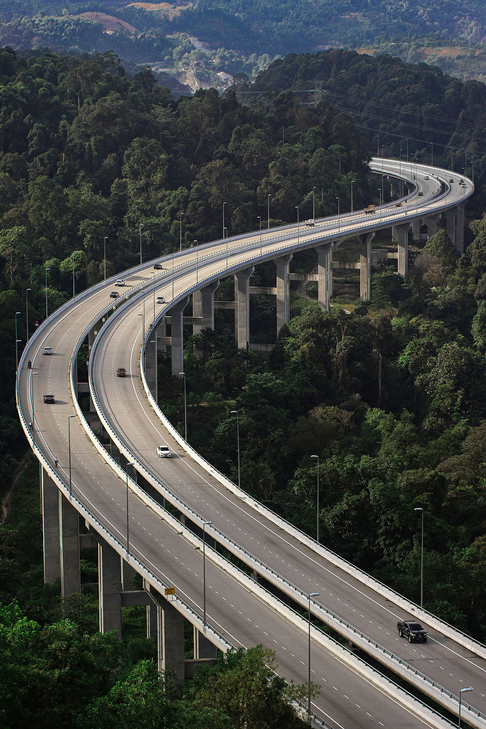 Curving elevated highway with a few cars crossing dense green rainforest hills under hazy mountains. Demonstrates a unique architectural design and how our freeway and highway systems can vary throughout different landscapes.