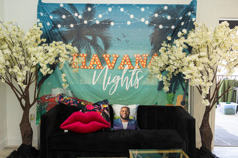 Havana nights backdrop behind black couch with a custom pillow. A white blossom tree sits on both sides of the black couch.