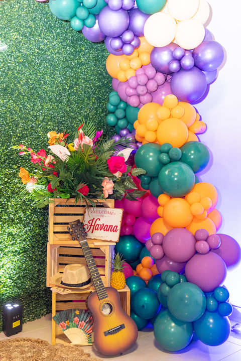 A close up of tropical-colored balloons in front of a grass wall backdrop. Three stacked crates, a ukulele, sun hat, fan and Havana sign sit next to the balloons. Photo by Ideal Shots LLC.