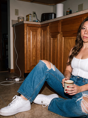 Krista sitting on a kitchen floor with a coffee mug, photographed with direct flash in a candid editorial portrait.