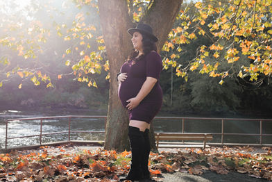 Pregnant woman in a maroon dress poses outside with fall foliage and a river.