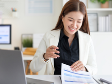 Smiling woman in white blazer reviews financial statements beside a laptop in a bright office, with HFM logo on screen.