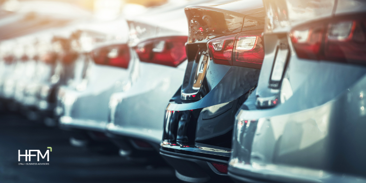 Row of new cars parked closely, back view with red taillights. Sunlit, sleek surfaces. HFM logo visible on the left.