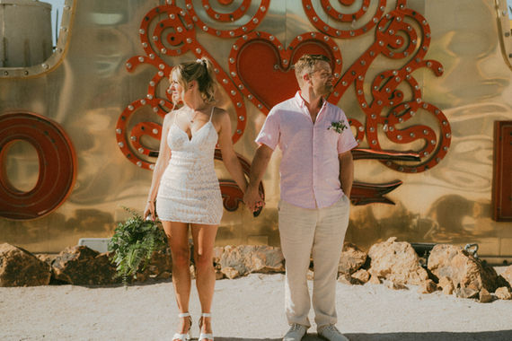 Couple holds hands in front of heart-shaped neon with wedding attire.