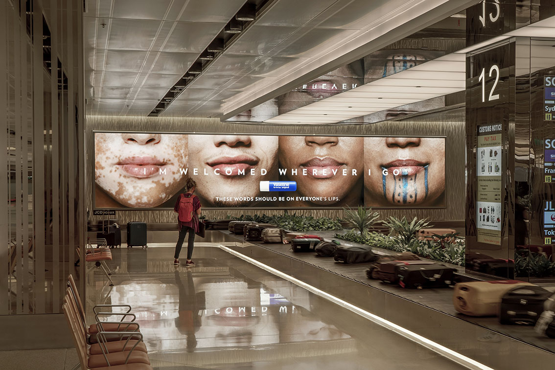 Airport baggage claim with a large Vaseline ad showing faces and the text I'M WELCOMED WHEREVER I GO; one traveler stands nearby.