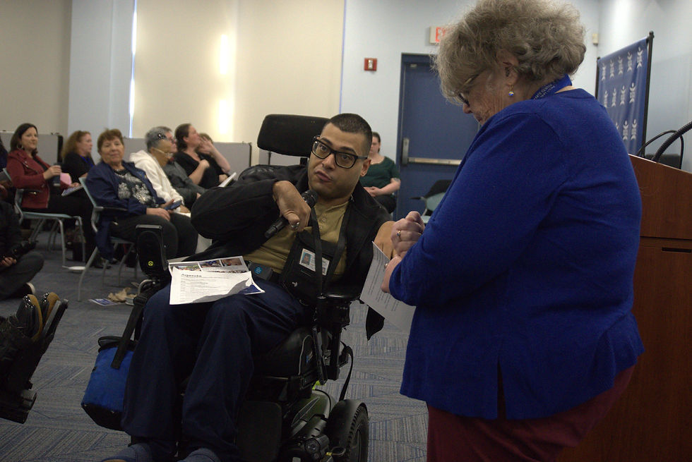 Myles Husband-Brawn, a member of the Peer Support & Advocacy Group, speaking to an audience from his wheelchair while a colleague assists at the podium.