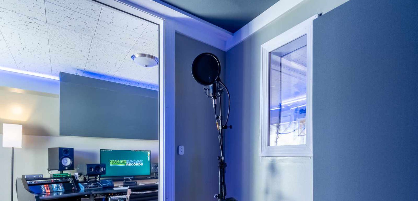 View from the Studio B isolation booth into the control room, with a mic, pop filter, and headphones in the foreground