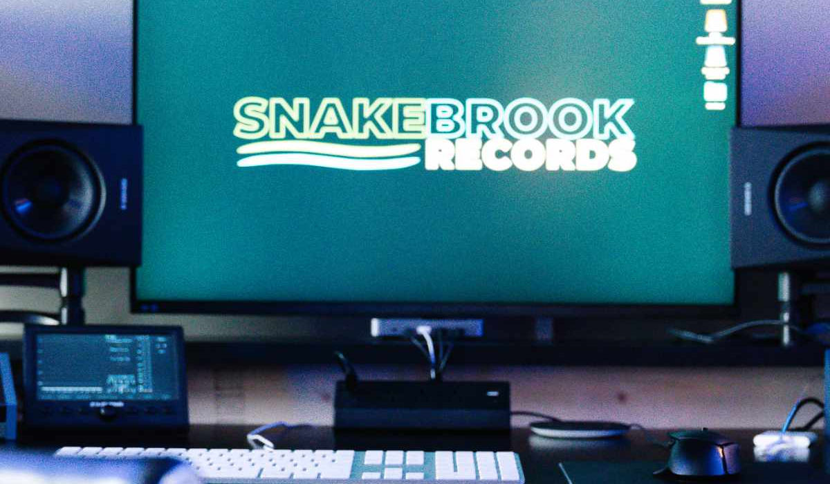 Studio B at Snake Brook Records in Natick, MA, with a large screen showing the studio logo, flanked by studio monitors above a MIDI keyboard and desk chair.